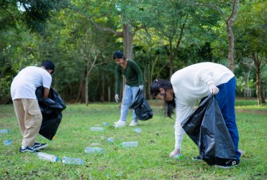 Bir grup kendini adamış genç gönüllü parktaki bir çevre temizleme etkinliğine katılıyor. Gönüllüler büyük siyah çantalar taşıyor. İnsanlar parkta çöp ve plastik şişe topluyor..