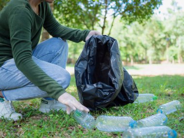 Gönüllü bir kadın parktaki bir çevre temizleme etkinliğine katılıyor. Eldiven takan ve büyük siyah çantalar tutan bir kadın. Parkta çöp ve plastik şişe toplayan bir kadın..