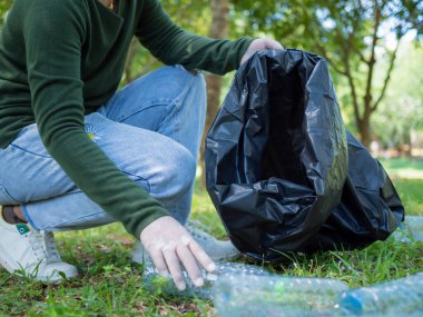 Gönüllü bir kadın parktaki bir çevre temizleme etkinliğine katılıyor. Eldiven takan ve büyük siyah çantalar tutan bir kadın. Parkta çöp ve plastik şişe toplayan bir kadın..