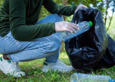 Gönüllü bir kadın parktaki bir çevre temizleme etkinliğine katılıyor. Eldiven takan ve büyük siyah çantalar tutan bir kadın. Parkta çöp ve plastik şişe toplayan bir kadın..