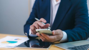 İş dünyasında mobil verimliliği, teknolojiyi ve çevrimiçi veri yönetimini temsil eden, ofis masasında dijital iletişim ya da finansal analiz için akıllı telefon kullanan bir işadamının yakın çekimi.