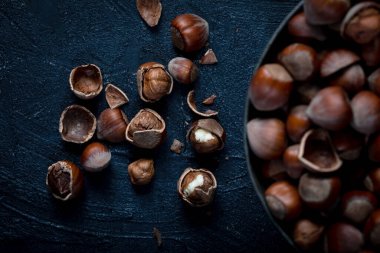 Cracked and unbroken shelled hazelnuts with a bowl of hazelnuts on a dark background.