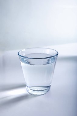 Su dolu berrak bir bardak pürüzsüz, beyaz bir yüzeye dayanır.. 