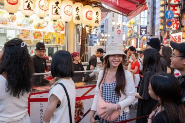 Japonya 'nın işlek Dotonbori semtindeki takoyaki sokak yiyecek standında sıra bekleyen insanlar. Sahne, Japon gece hayatının canlı atmosferini parlak fenerler, işaretler ve çeşitli yerel ve turist gruplarıyla yakalıyor. Haziran 2025.