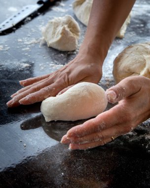 Yakın plan bir el hamur parçasını koyu renkli bir tezgahta küçük bir somun haline getiriyor. Arka planda başka hamur parçaları ve büyük bir mutfak bıçağı var.. 