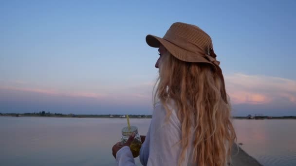 白いシャツと帽子の長い白い髪の美しい女の子は 日没時に海を見ています 海岸でのリラクゼーションの概念 恋の女の子は夕日を見て — ストック動画