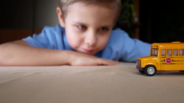 可愛い男の子がおもちゃの黄色いスクールバスをテーブルの上に転がしています 距離学習中に学校を逃す子 — ストック動画