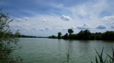Manzara. Mavi göl suyu, üstünde beyaz bulutlar. Gölün etrafındaki yeşil ağaçlar. Vahşi doğada yolculuk. Yazın büyük gölün üzerinde bulutlu mavi gökyüzü