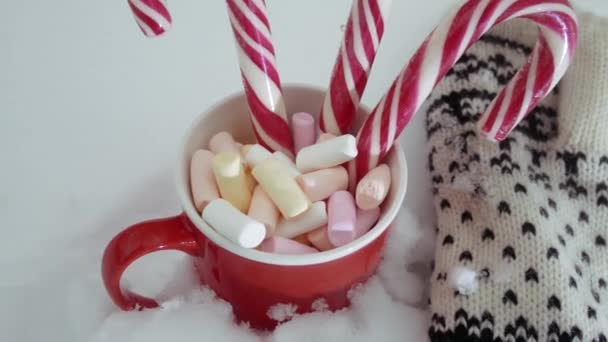 白い雪を背景に 赤いカップにマシュマロとクリスマスの赤と白のロリポップ 隣には伝統的な装飾が施されたミトンがあります — ストック動画