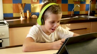 A cute boy of 7-8 years old in headphones sits in the kitchen in front of the tablet and talks to someone from the screen. Online lessons, distance online learning. Communication and teaching children