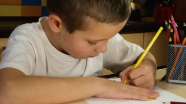 A cute boy of 6-7 years old sits at a table in a room and draws a drawing with a yellow pencil. Child development. Teaching children. Distance learning for children
