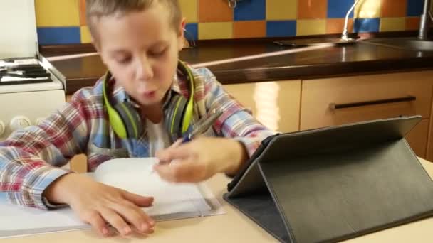 首の周りにヘッドフォンを付けた再生シャツを着た6 7歳の少年がキッチンのテーブルに座り タブレットの画面を見て ノートにペンで書きます 距離学習 オンライン — ストック動画