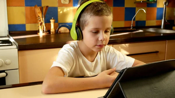 A boy of 6-7-8 years old sits in the kitchen in a white T-shirt and bright green headphones on his head, looks at the tablet screen, listens and answers. The concept of online learning, communication