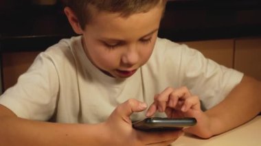 A cute boy sits in a room and holds a smartphone in his hands. Scrolls your finger across the screen. A child plays an online game on his smartphone. The child communicates in social networks using a