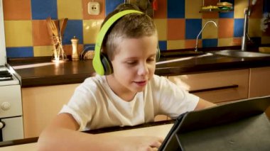 A boy of 6-7-8 years old sits in the kitchen in a white T-shirt and bright green headphones on his head, looks at the tablet screen, listens and answers. The concept of online learning, communication