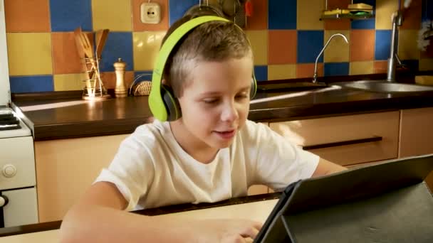 8歳の男の子は 白いTシャツと頭の上の明るい緑のヘッドフォンでキッチンに座って タブレット画面を見て 聞いて答えを聞きます オンライン学習 コミュニケーションの概念 — ストック動画