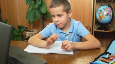 A 7-year-old schoolboy writes and shows a notebook to the monitor to the teacher. Remote homeschooling during the pandemic