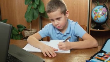 A cute boy of 7-8 years old sits in front of a laptop in his room during an online lesson. The boy writes with a pen in a notebook, shows the notebook in front of the monitor. Answers questions