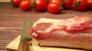 A piece of bacon with meat, pork fat, on a cutting board on the table. In the background there are tomatoes. Cooking meat in the kitchen