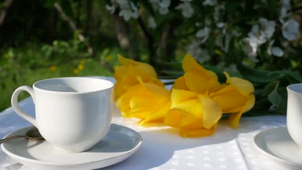 白いティーカップとソーサー 黄色のチューリップと庭のテーブルの上のケーキの作品 春の屋外ランチ — ストック動画