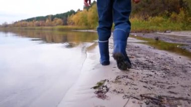 Childs feet in rubber boots walk along the lake shore in autumn. In the background a lake and yellowing trees. Walk along the lake in autumn