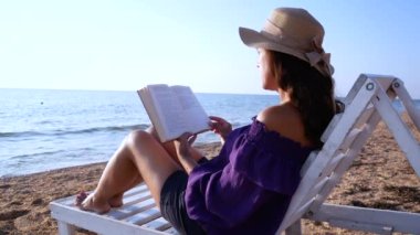 A girl in a straw hat sits on a white sun lounger on the beach with a book in her hands. A calm blue sea ahead