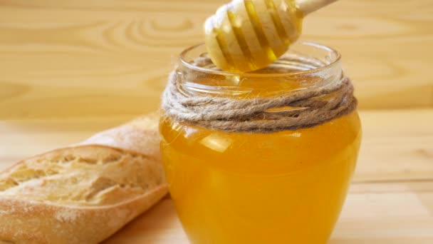 一只木制勺子从玻璃瓶中盛放金黄色蜂蜜的液体 附近躺着一个小面包 有机蜂蜜 天然产品和适当营养 — 图库视频影像