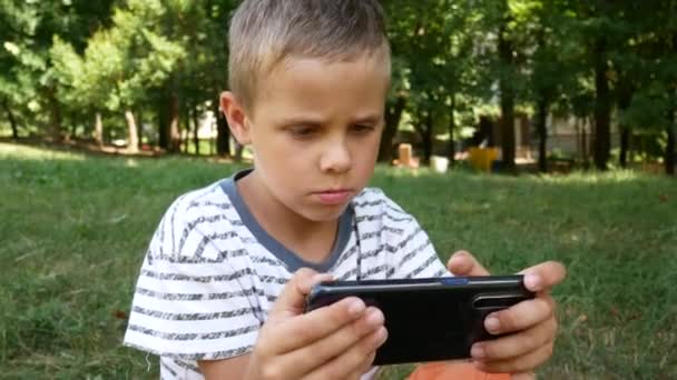 熱意を持って6 7歳の少年は スマートフォンの画面を見て 携帯電話上でゲームをプレイします 背景の公園 緑の木 — ストック動画
