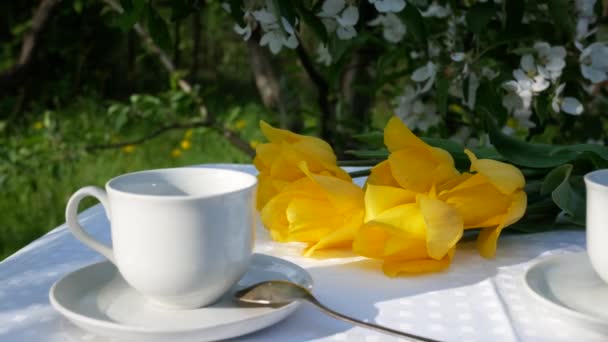 ライフスタイル 庭でお茶を飲む 庭のティーテーブルの上にカップ スプーン スライスされたマフィン テーブルの上の背景には黄色のチューリップの花束があります — ストック動画
