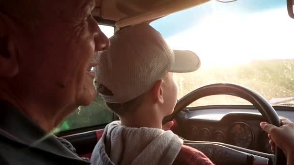 少年と彼の祖父は 一緒に車を運転し 車の中でホイールを保持します 子供はハンドルを回す ウィンドウを通してフィールドが表示されます 太陽の光が車内を照らす — ストック動画