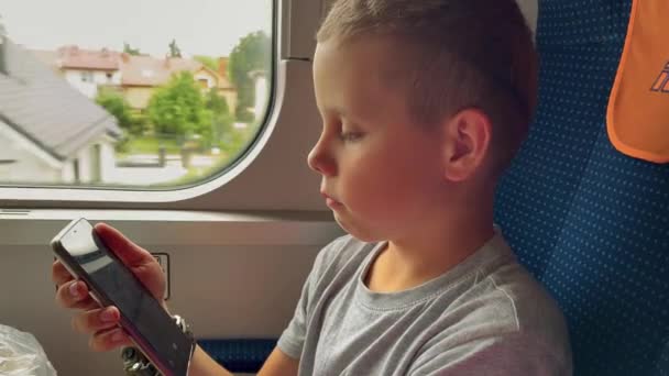 一个穿着灰色T恤的男孩坐在城际列车的一辆行驶中的车上 看着智能手机屏幕 你可以看到不断变化的风景和村庄的房屋 — 图库视频影像