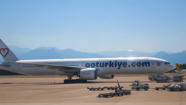 アンタルヤ トルコ 2023年6月25日 大きな飛行機が山を背景に滑走路に立っています 近くには荷物を積むための公共交通機関があります 大規模な旅客空港の仕事 — ストック動画