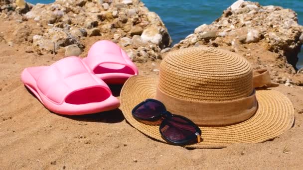 粉色海滩堵塞 草帽和太阳镜在沙滩上 背景是岩石和蓝色的海水 海滨度假的概念 — 图库视频影像