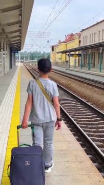 Bir çocuk tren istasyonundaki bir tren istasyonunda bavul taşıyor. Demiryolu platformunun yan tarafında istasyon binası var. Yaz tatillerinde seyahat ve gezi kavramı.