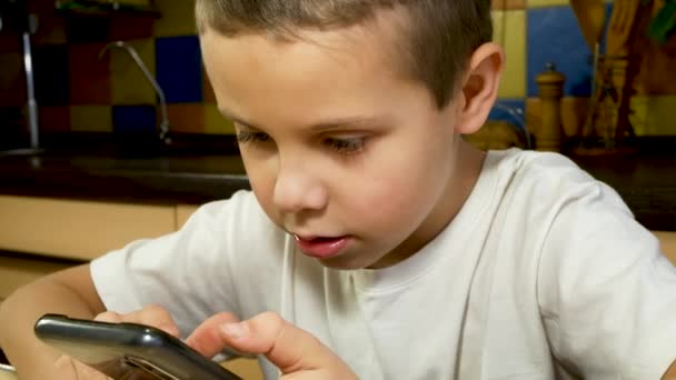 7歳の少年が台所で携帯電話を手にした 少年はスクリーンにスクロールし ガジェットの助けを借りてソーシャルネットワークでのコミュニケーション 子供とガジェット エレクトロニックゲーム — ストック動画