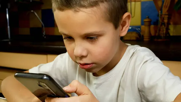 A boy of 6-7 years old with a phone in his hands in the kitchen. The boy scrolls on the screen, talks. Communication in social networks with the help of gadgets. Kids and gadgets. Electronic games.