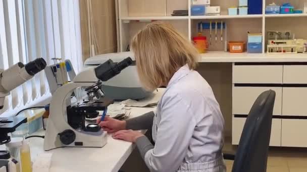 ウクライナのハリコフ2024年2月1日 女性実験アシスタントが顕微鏡を検査し 一枚の紙にデータを書き込む 医学研究所の研究 臨床血液カウント — ストック動画