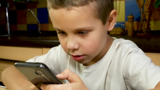 一个7 8岁的可爱男孩坐在那里 手里拿着智能手机 在社交网络上交流 儿童和小玩意 远程学习 智能手机游戏 — 图库视频影像