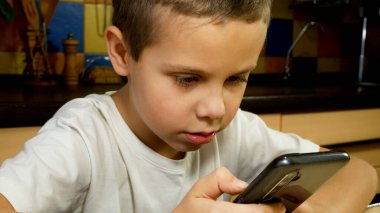 Mutfakta elinde telefon olan bir çocuk ekranı dinleyip konuşuyor. Cihazları ve internet uygulamalarını kullanarak iletişim. Cep telefonlarında çocuklar ve oyunlar.