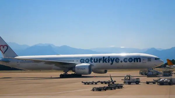 stock image Antalya, Turkey - June, 25, 2023: A plane on the runway at the airport of a tourist city. In the background are mountains. In front of the plane there are auxiliary cars with luggage trolleys. Baggage