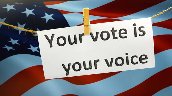 Рука вешает табличку с надписью "Ваш голос - ваш голос на фоне американского флага". Президентские выборы в США. Люди отдают свои голоса за кандидатов Камалу Харрис или Дональда