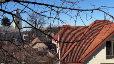 Açık mavi gökyüzünün arka planında ve Katolik kilisesinin kulesinde, ağaç dalları arasında kırmızı kiremitler olan çatı..