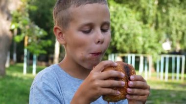 İştahlı bir çocuk yazın dışarıda hamburger yer. Arkasında yeşil bir park var. Çocuklar için fast food. Sağlıklı beslenme kavramı.