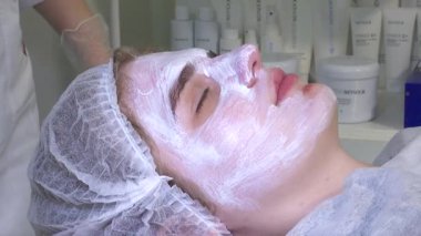 Genç bir kadın, yüzünde beyaz bir maskeyle güzellik muayenehanesinde yatıyor. Yüz bakımı konsepti. Kozmetik. Dikkat et, deriyi nemlendirir, kırışıklıklarla savaşır. bir kozmetikçide bakım.