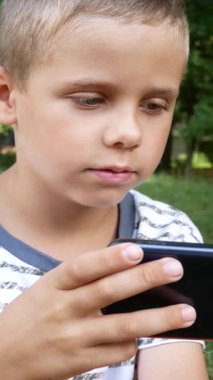 Çocuk 6-7-8 yaşında. Akıllı telefon ekranına bakıyor. Parktaki cep telefonuyla oyun oynuyor.