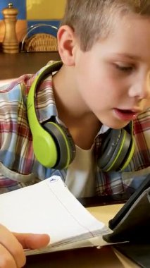 A boy of 6-7 years old in a plaid shirt with headphones around his neck sits at a table in the kitchen, looks at the tablet screen and writes with a pen in a notebook. Distance learning. Online