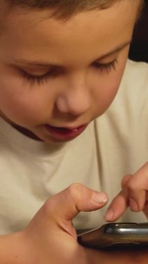 A cute boy sits in a room and holds a smartphone in his hands. Scrolls your finger across the screen. A child plays an online game on his smartphone. The child communicates in social networks using a