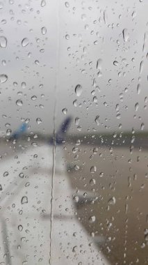 Yağmur damlalarının aktığı bir pencere. Arka planda, uçağın kanadı pencereden görülebilir. Yağmurda uçak uçurmak. Uçmayan hava.