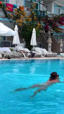 Side, Türkiye - 17 Haziran 2023: Bir kız Akdeniz tatil köyündeki bir otelin mavi su havuzunda yüzüyor. Kenarlarında beyaz şemsiyeli güneş şezlongları var. Otelde dinlenmek için..