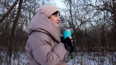 Ilık ceketli genç bir kadın, karlı bir kış parkında termostan sıcak çay içiyor. Noel havası, kış yürüyüşü, rahat bir an..